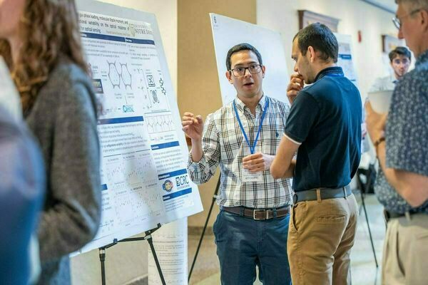 A man in a plaid shirt gestures while explaining his research poster to another man at a University of Notre Dame event.  A third person looks on.  The poster includes scientific charts, diagrams, and text.