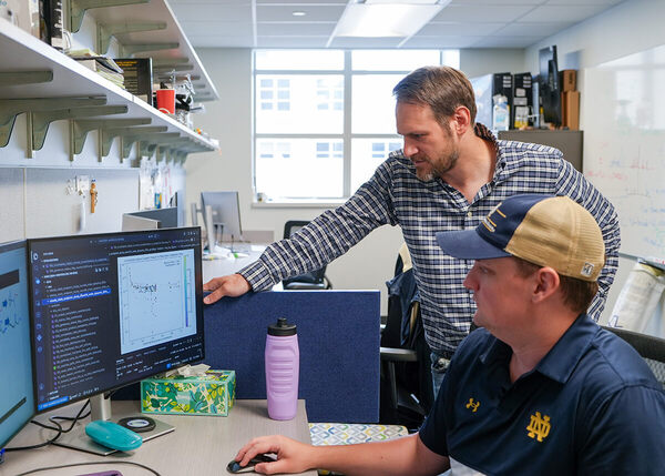 Two men examine a computer monitor displaying a data plot in an office. One wears a Notre Dame polo, the other a plaid shirt.
