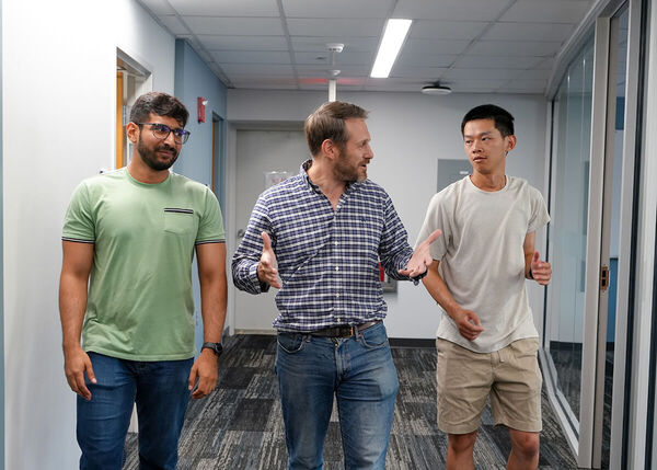 A bearded man in a blue plaid shirt gestures as he speaks to two younger men walking with him in a hallway.
