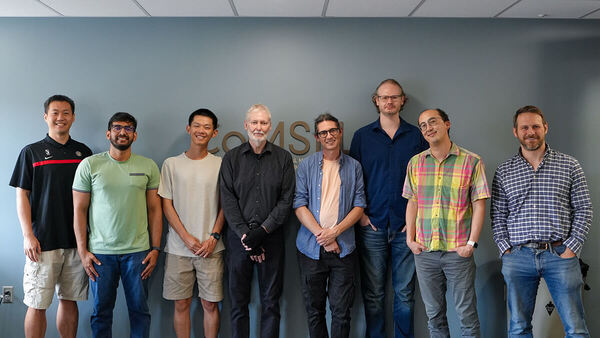 Eight diverse men, some with beards or glasses, stand smiling in casual clothes against a blue-gray wall.